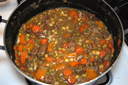 A bowl of soup with meat and vegetables cooking on a stove, with Kitchen and Stew