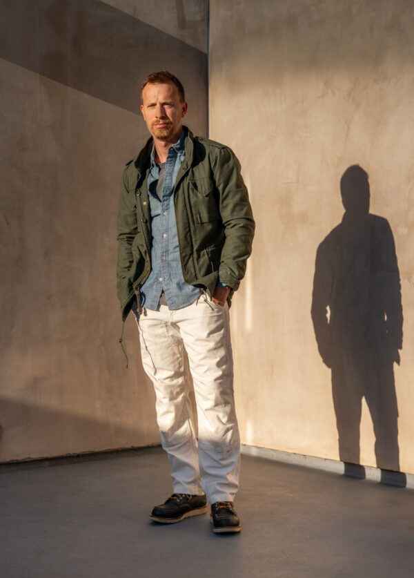 a man wearing a field jacket over chambray shirt and slim fit pants