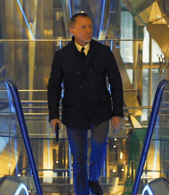 daniel craig james bond wearing a peacoat over a shirt and tie, with slim fit pants, and coming up an escalator holding a firearm