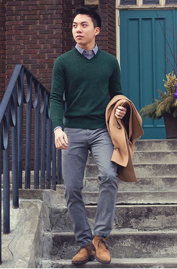 a man wearing fashion business casual outfit with green v-neck sweater, dress shirt, grey dress pants, and chukka boots with an overcoat draped over his arm