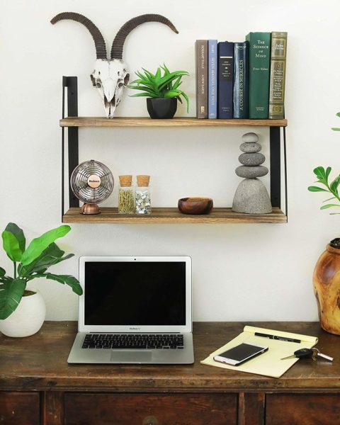 a space saving floating bookshelf on a wall holding books and small home decor items, in a workspace area with a laptop, mobile phone, writing pad, and faux plants