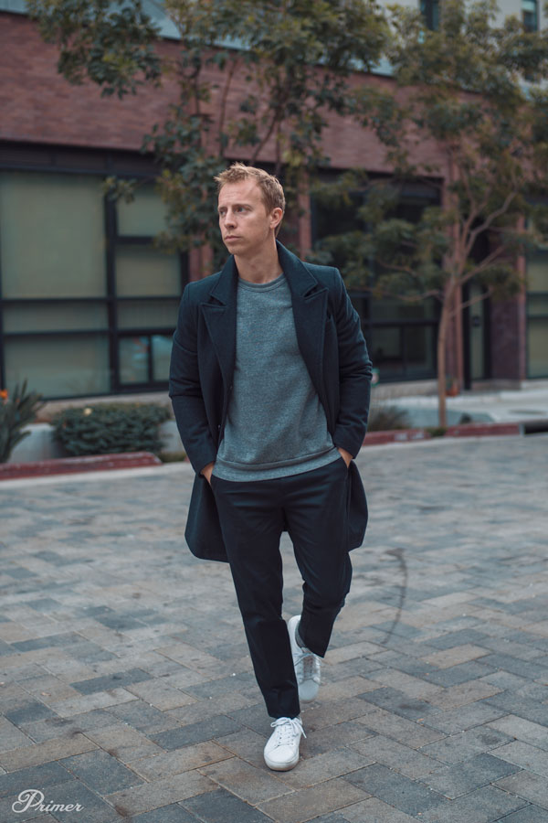 a man wearing a smart casual outfit of a topcoat, gray sweatshirt, black pants, and white sneakers