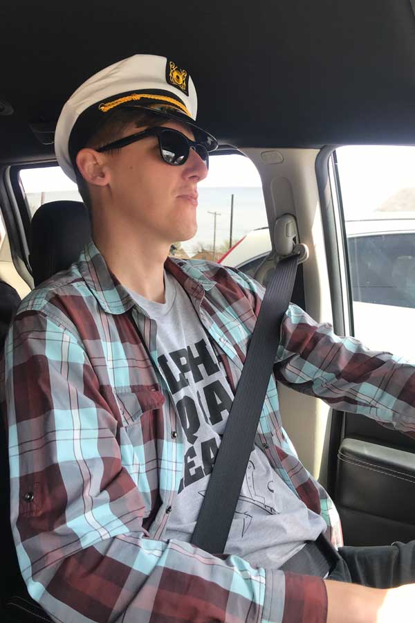 a man in the driver's seat of a car wearing a captain style hat, sunglasses, and a casual shirt