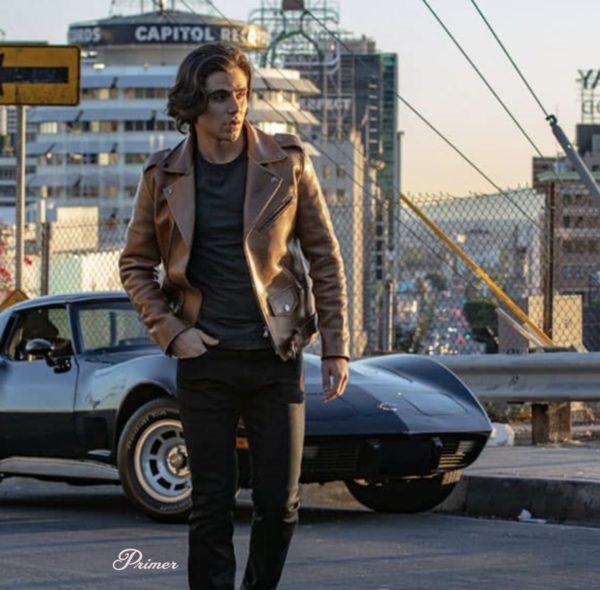 a man wearing a brown leather motorcyle style jacket over a shirt and slim jeans, with a parked Corvette and city buildings in the background