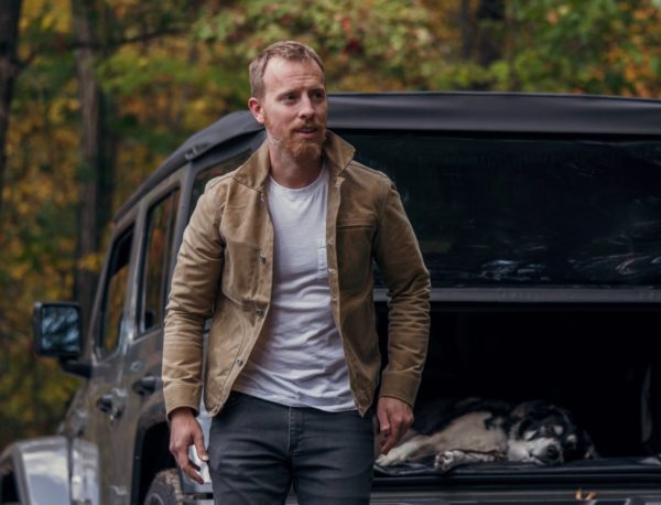 a man wearing a waxed canvas jacket over a shirt and jeans, with a relaxing dog in the rear compartment of a vehicle in the background