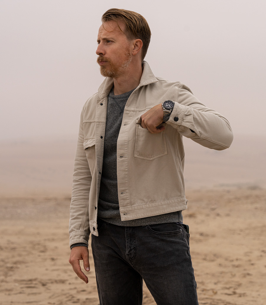 Man wearing G-Shock MTG Watch with tan trucker jacket, grey sweatshirt, and faded black jeans in desert