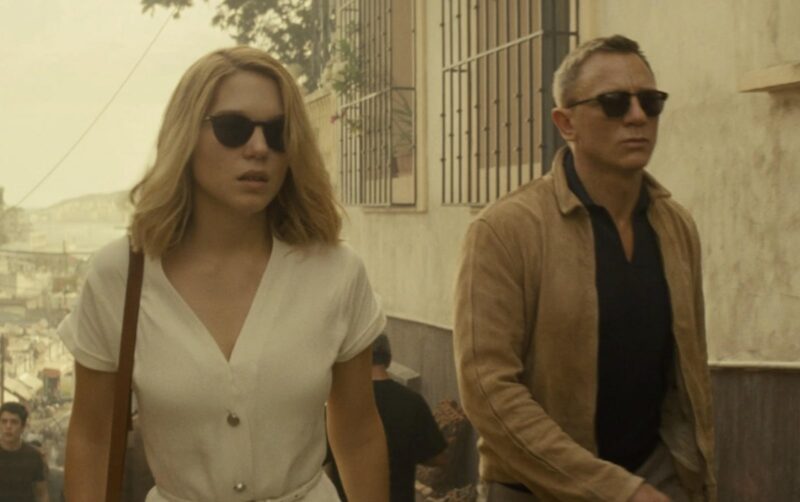 daniel craig james bond wearing a waxed jacket, collared shirt, and sunglasses, walking alongside a woman wearing sunglasses and a dress