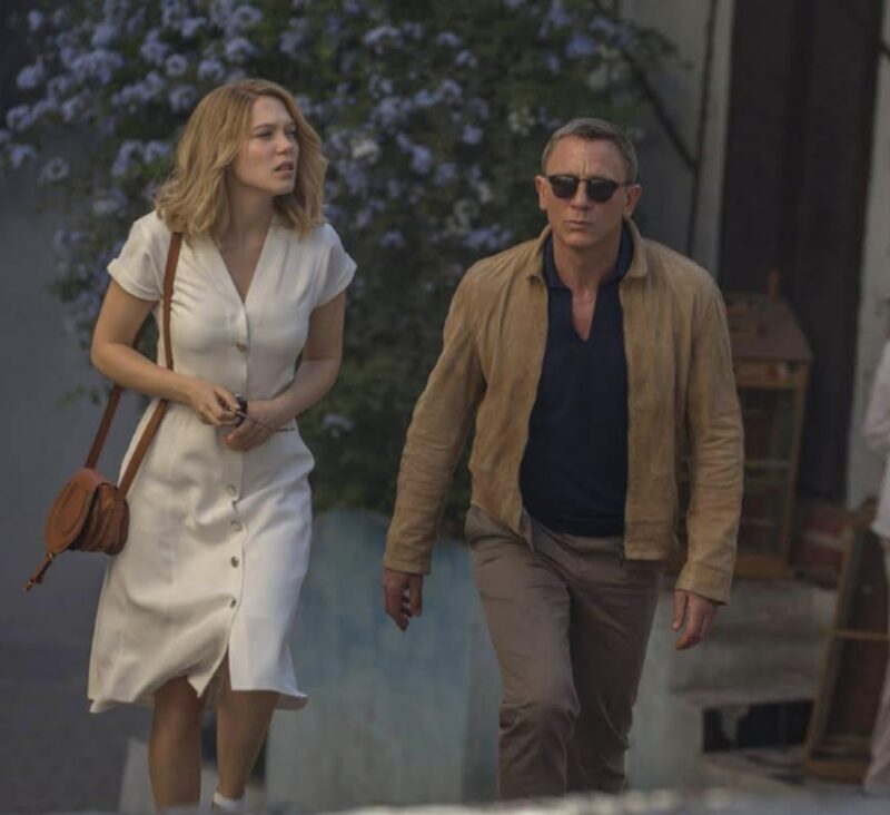 daniel craig james bone wearing a waxed jacket, collared shirt, and pants, walking alonside a woman in a long white dress