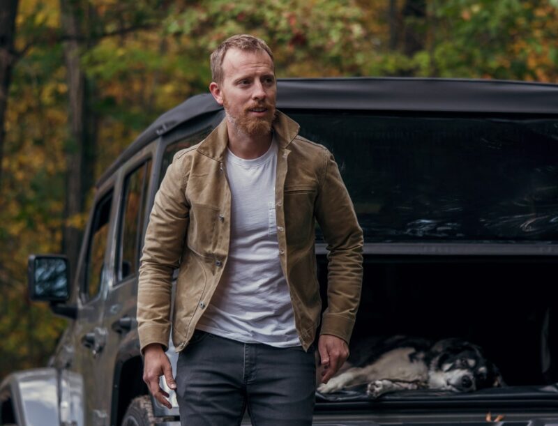 a man wearing a waxed jacket over a shirt and jeans with a relaxing dog in the rear compartment of a vehicle in the background