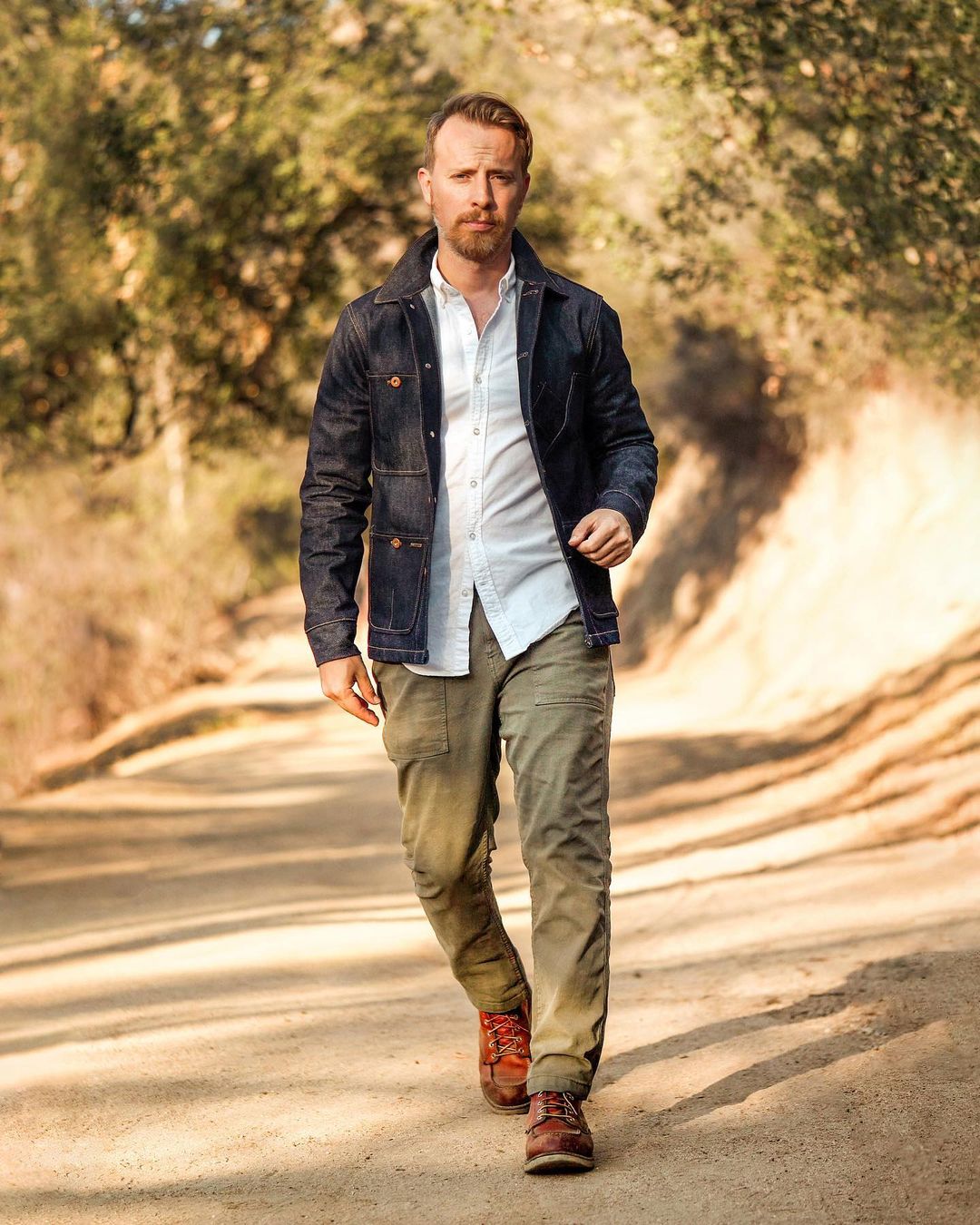 man wearing a denim tellason chore coat with white oxford shirt, green og 107 pants and red wing boots