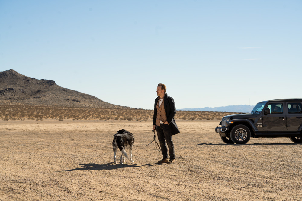 pria berdiri di gurun dengan anjing dan jip di latar belakang