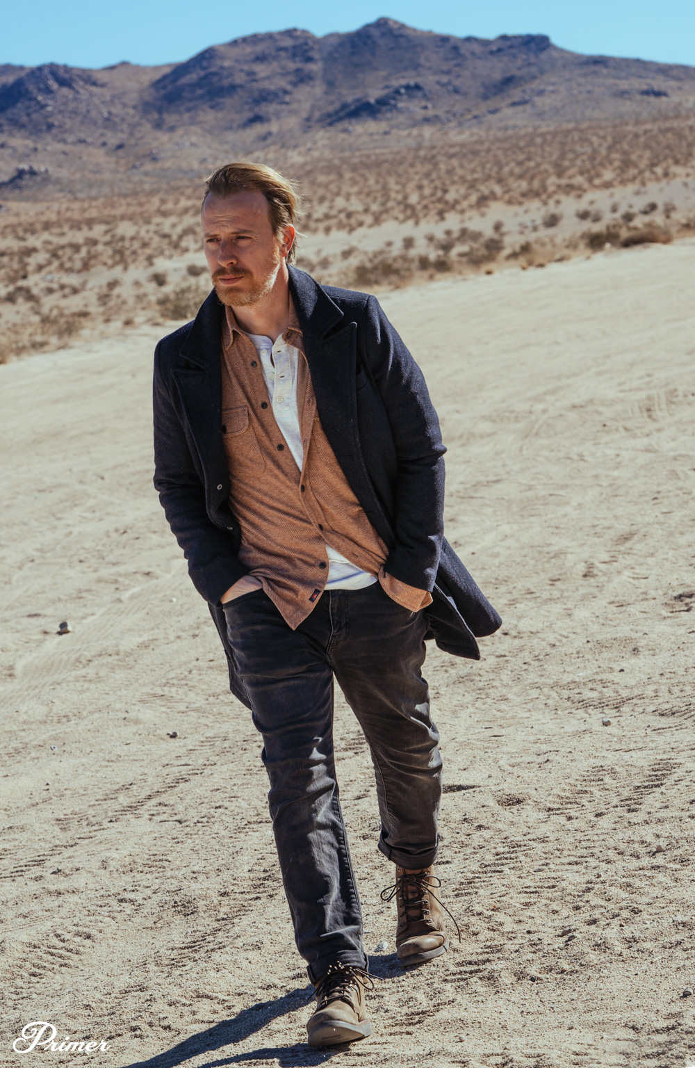 Andrew Snavely wearing a casual outfit in the desert with a topcoat, fatherly legend sweater shirt, cloud henley, gray denim, and brown Chippewa boots