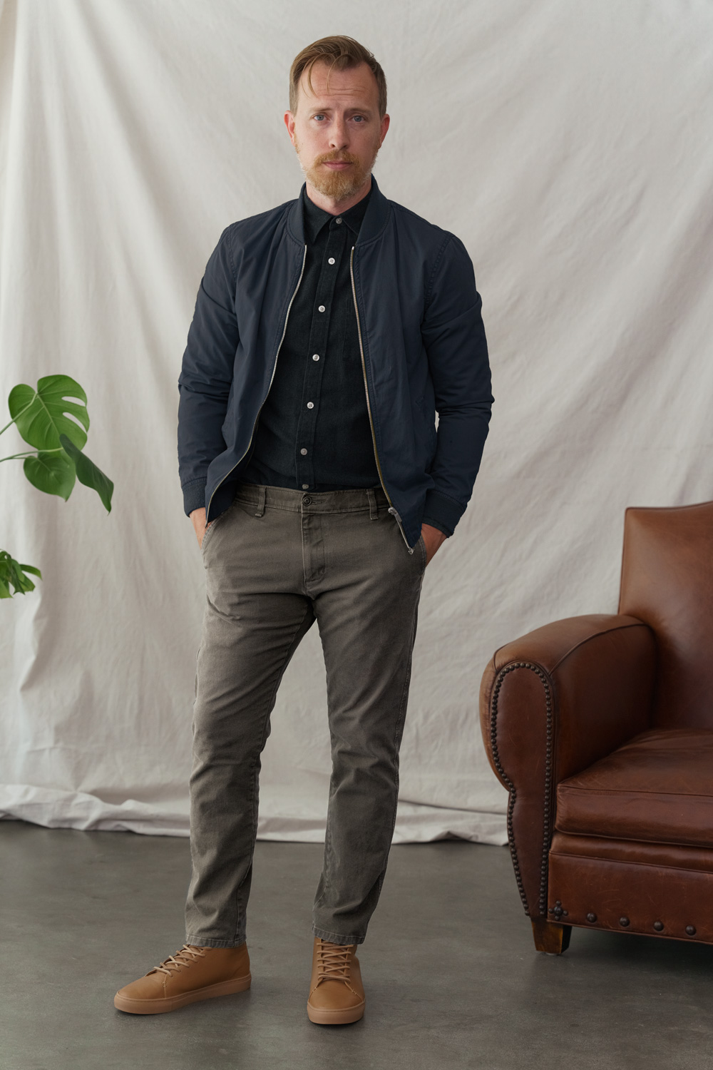 a man wearing a smart casual outfit featuring blue bomber jacket, black button up shirt, chinos, and tan high top sneakers