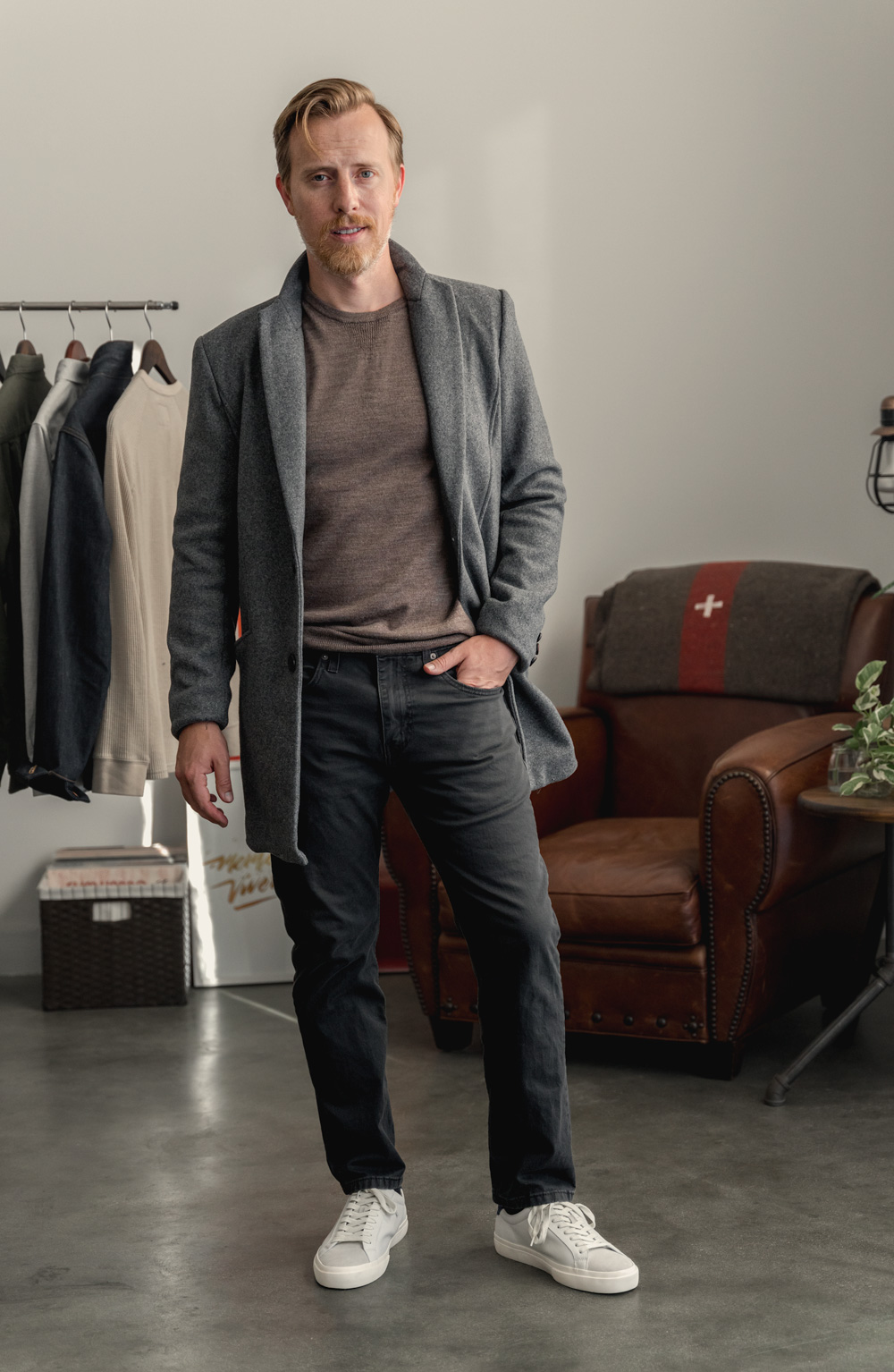 a minimalist men's outfit with gray topcoat, brown sweater, dark gray jeans, and white sneakers