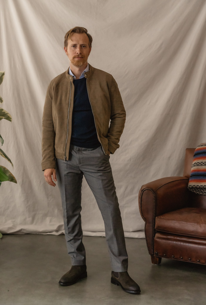 a suede leather jacket worn with a business casual outfit consisting of a navy sweater, blue dress shirt, gray trousers and olive suede chelsea boots