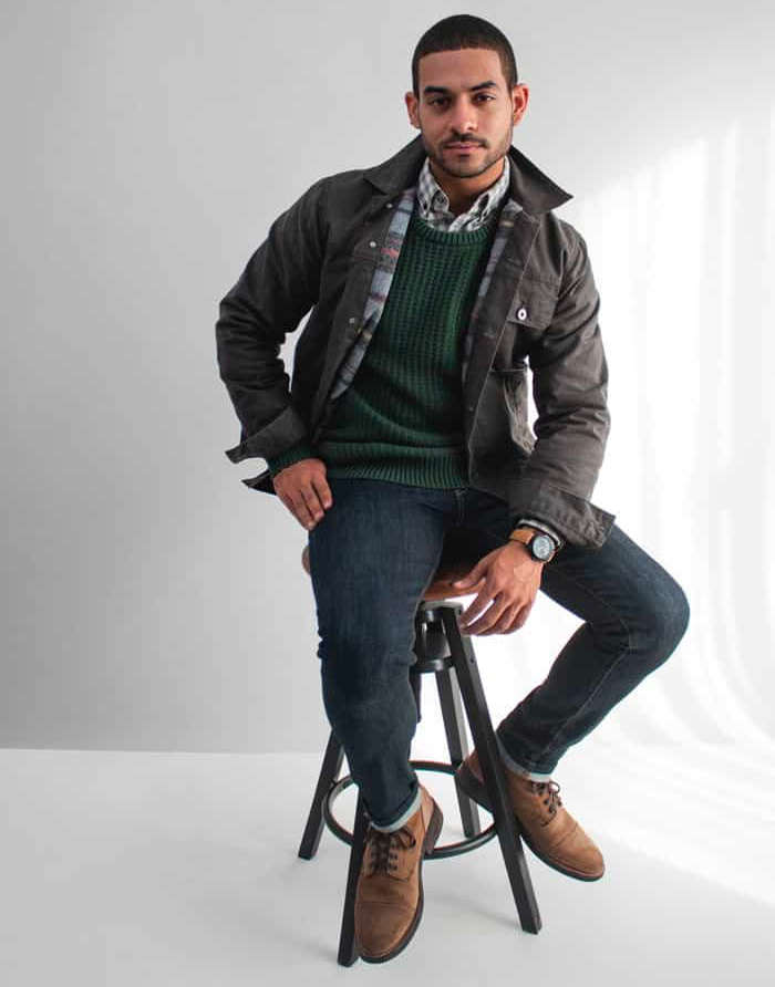 waxed canvas jacket prep outfit with green sweater, plaid shirt, blue jeans, and tan boots