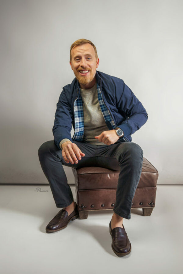 a man sitting on an ottoman wearing a harrington style jacket with pants and loafers