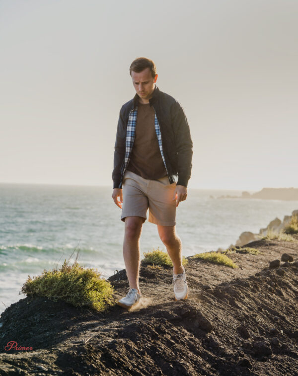 a man wearing a harrington style jacket over a shirt and shorts with sneakers walking along a coastline
