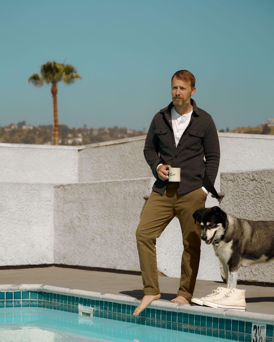 a man wearing khaki pants and a cardigan standing with a dog around a swimming pool