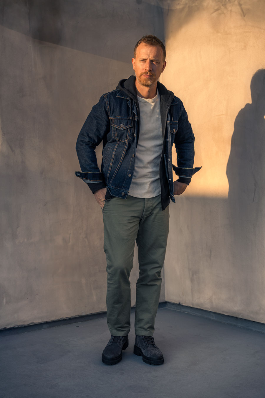 casual fall outfit for men featuring blue denim jacket, black hoodie, white thermal, green chino pants, and gray suede boots