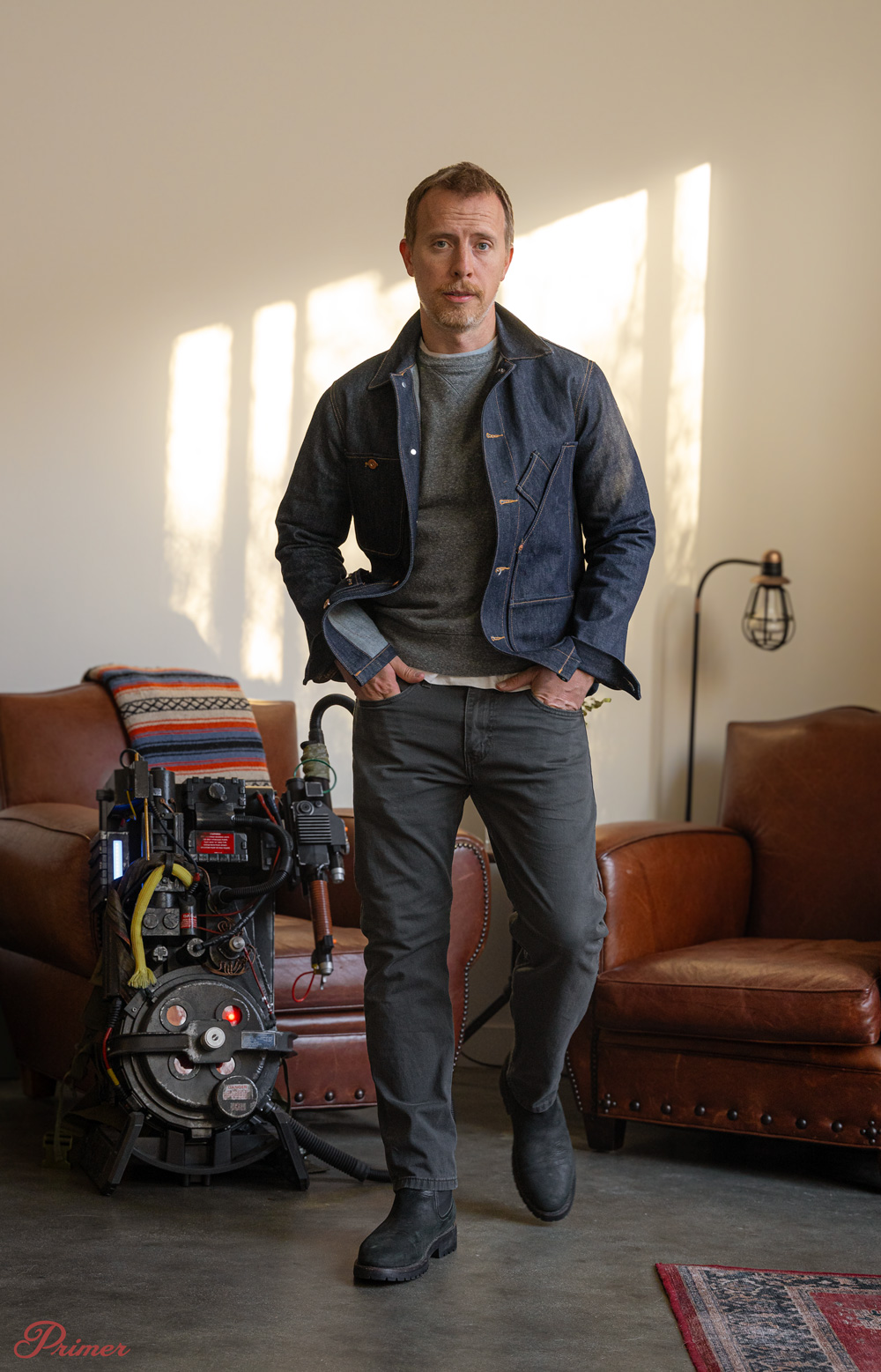 a man wearing a denim chore coat over a grey sweatshirt with charcoal pants and black boots in front of a proton pack from Ghostbusters
