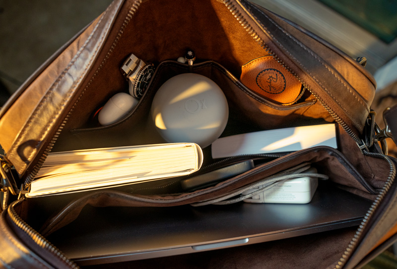 Interior view of an open Thursday Boots leather bag illuminated by warm light, showcasing various items including a notebook, a wristwatch, a white round case marked 'OX', a leather pouch with an embossed symbol, and electronic devices
