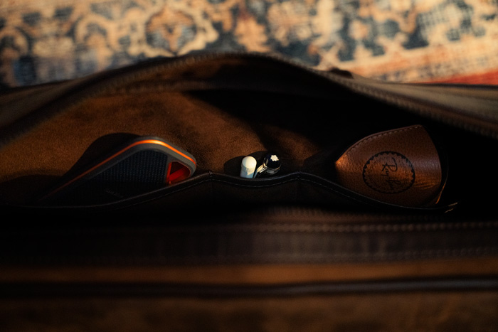 A close-up view inside a brown thursday boot co leather messenger bag reveals 3 pockets with a ssd, 2 pens, and a leather pouch, all set against the backdrop of a patterned rug.
