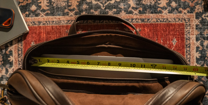 A leather bag open on a patterned rug, with a yellow measuring tape stretched across its width showing a measurement of approximately 14 inches