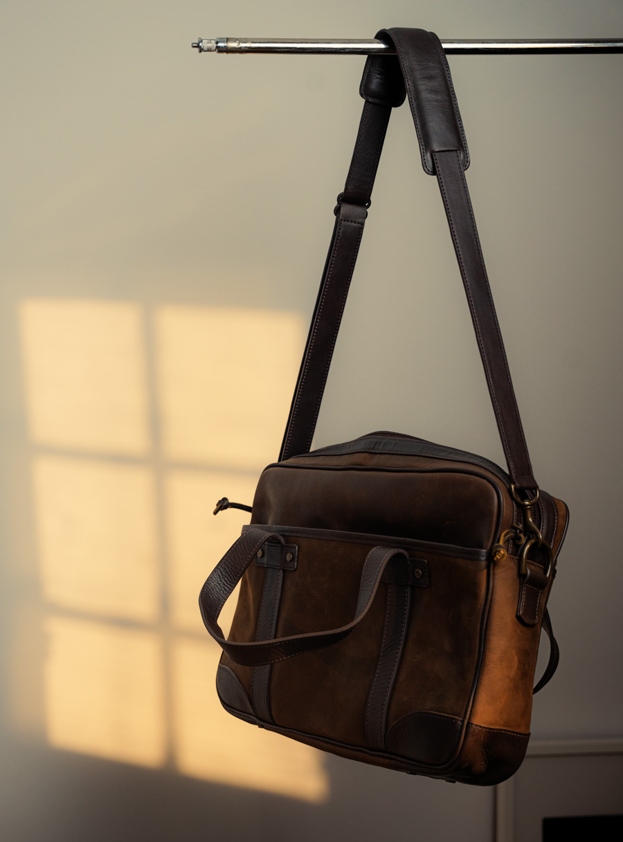 Brown leather messenger bag suspended by a leather shoulder strap from a metal stand, bathed in soft light with shadow patterns on the background
