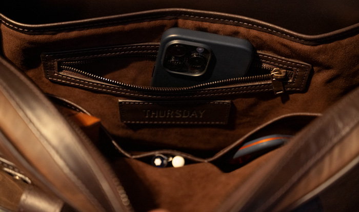 Close-up view inside a leather bag showing an interior pocket with a label reading 'THURSDAY', a smartphone, and other miscellaneous items partially visible