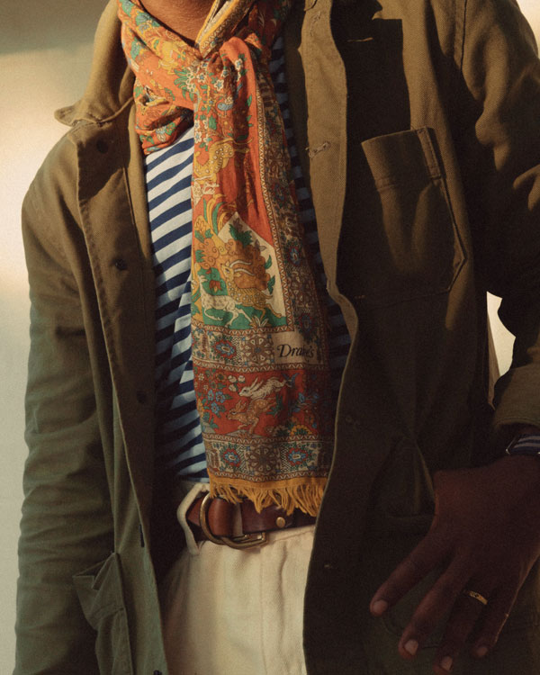 Close-up of a man's torso showing layered clothing and accessories. The person wears a blue and white striped shirt under an olive green coat, accented with a colorful scarf featuring red, blue, and yellow patterns. A brown leather belt is visible around the waist of white trousers. The lighting gives a warm tone to the image, and only the person's clothed midsection is visible; their face and lower body are not in the frame.