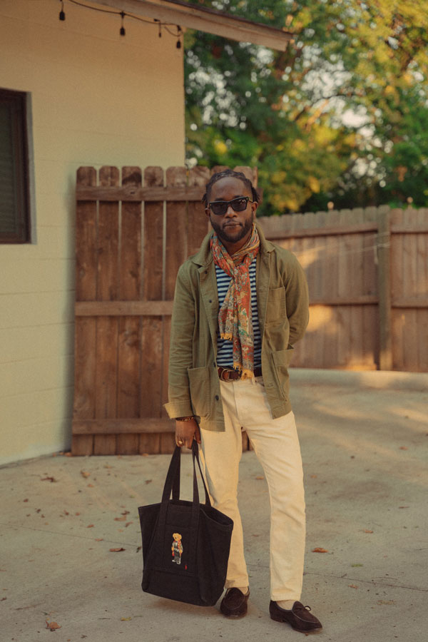 man with a unique personal style featuring a green coat, multi color scarf, striped tshirt, white pants, and character tote