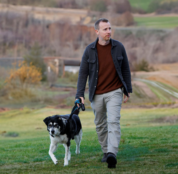 man walking his dog in a field, he's wearing a charcoal shirt jacket, burnt orange sweater, tan chino pants, and gray suede boots