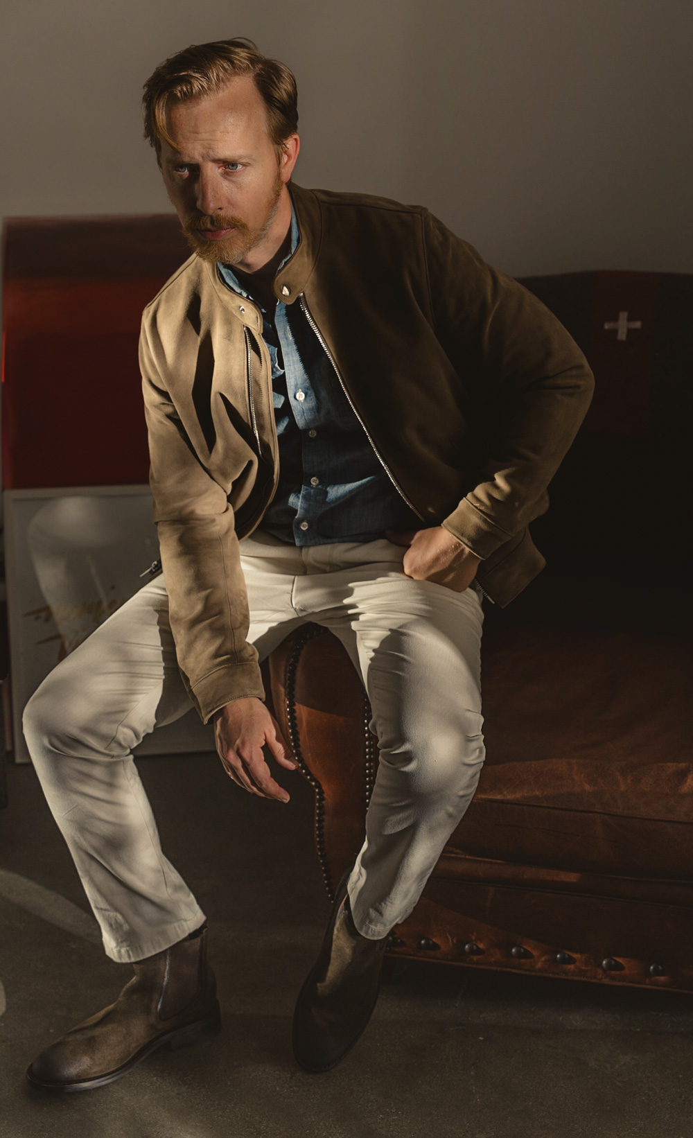 A man in a smart casual outfit sporting a brown bomber jacket over a denim shirt, paired with white pants that provide a sharp contrast to his brown boots.