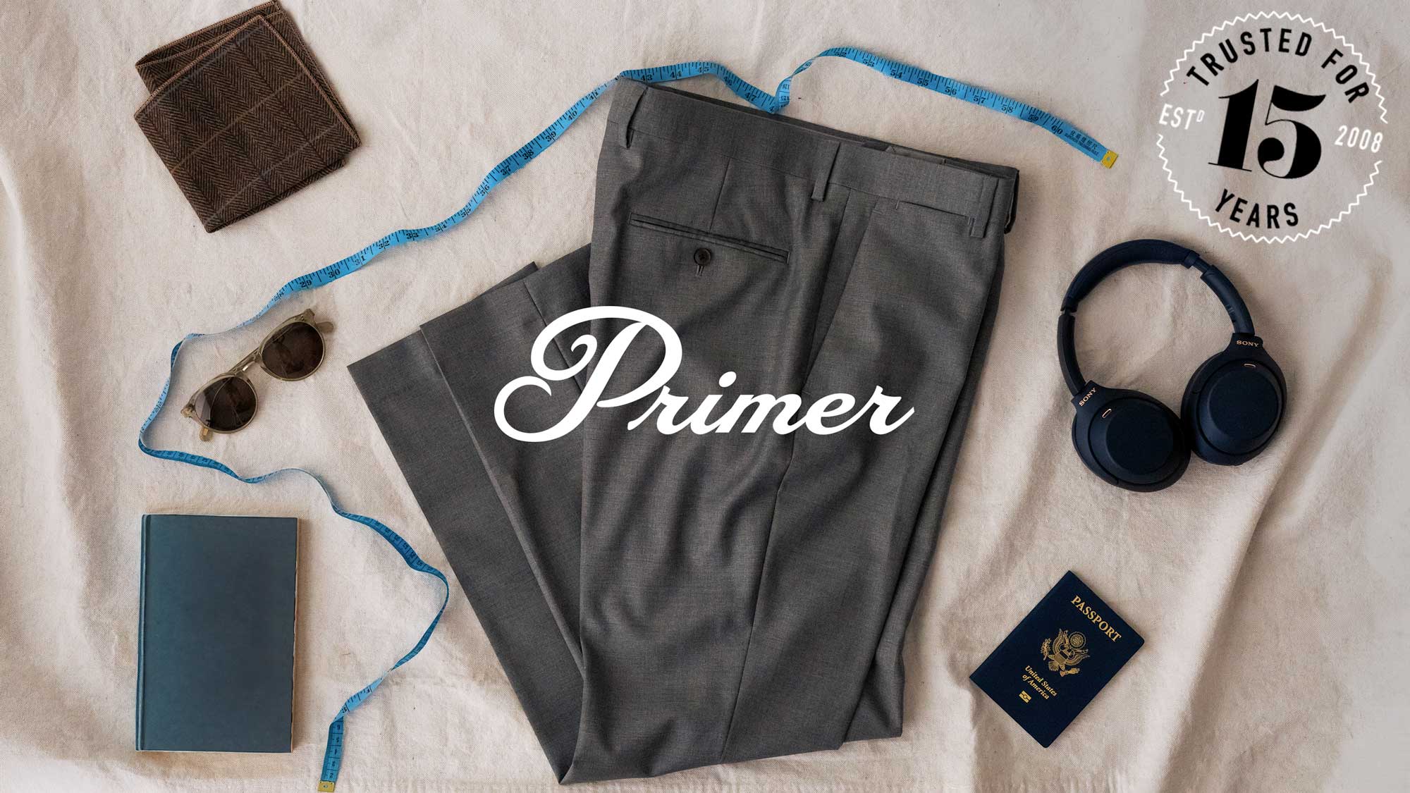 The primer logo and a badge that reads "trusted for 15 years, established 2008" over a photo of several items on a canvas backdrop – gray dress pants, a passport, wireless headphones, a journal, a measuring tape, sunglasses, and a pocket square