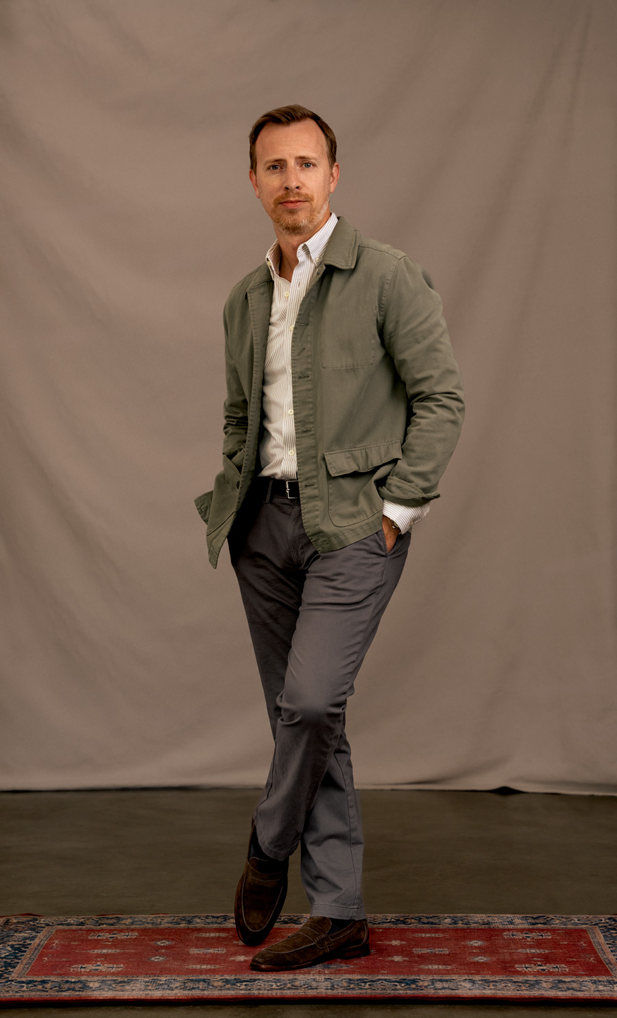 A man wearing an olive green chore coat as a blazer alternative on a business casual outfit featuring a white striped dress shirt, gray chino khakis, and suede loafers