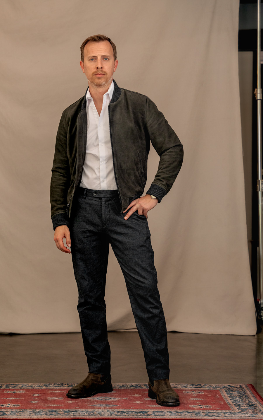 a man's outfit example using a suede bomber jacket as an alternative to a blazer. the outfit includes a white dress shirt, gray pants, and brown chelsea boots