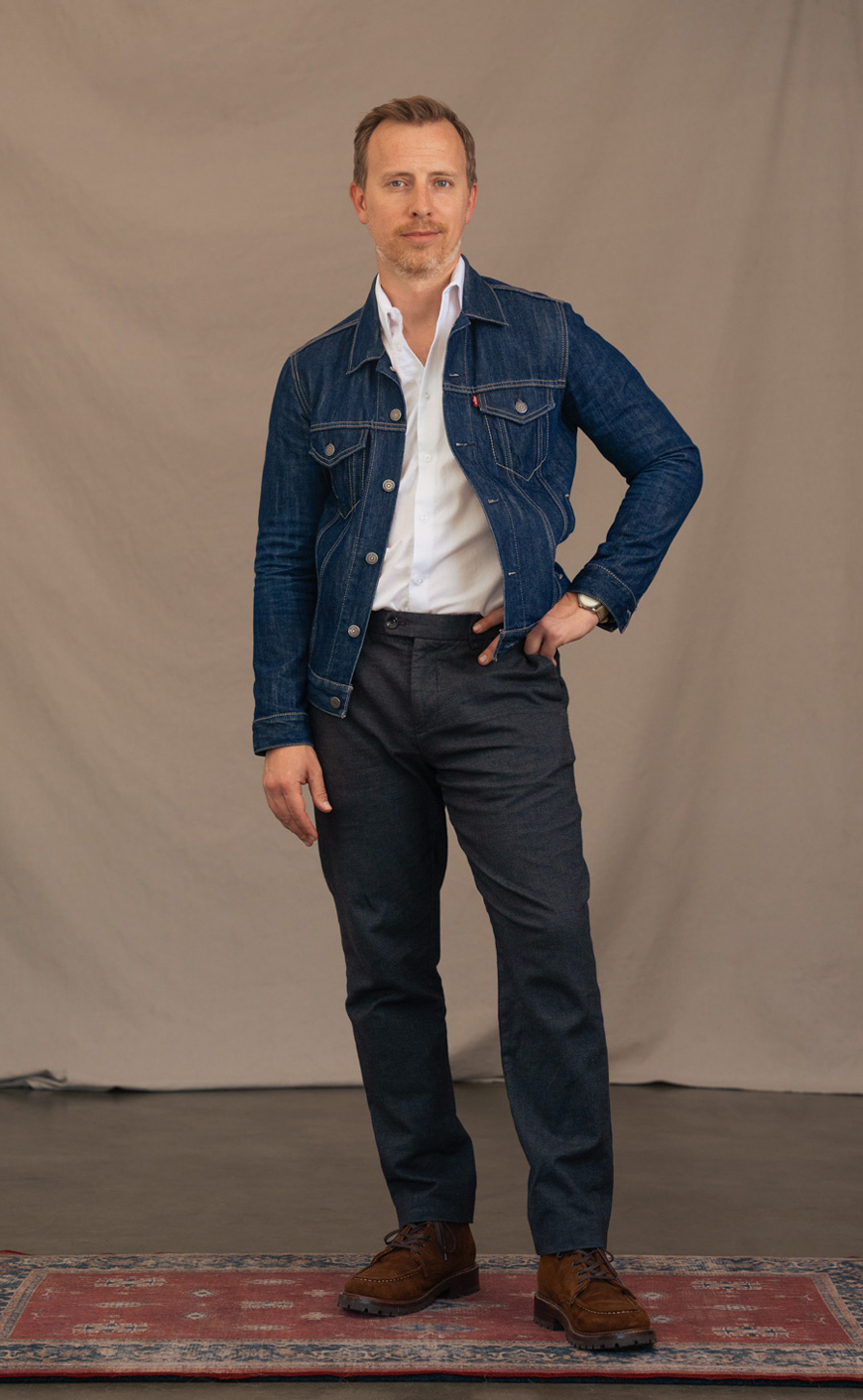 a men's outfit featuring a denim trucker jacket, white dress shirt, dark gray dress pants, and brown suede boots