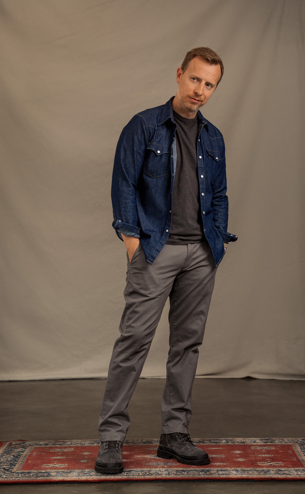 A man wears a dark grey t-shirt under an unbuttoned dark blue denim shirt  and chest pockets over a black pocket t-shirt. His outfit is completed with grey trousers and dark grey suede boots. He has short blonde hair, and he poses with one hand in his pocket.