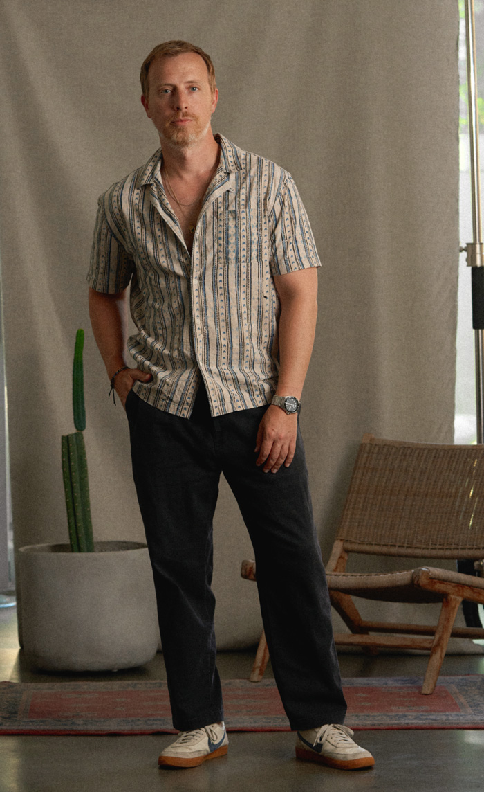 A man stands indoors against a neutral backdrop, wearing a short-sleeved, button-up shirt with a vertical striped pattern in beige and blue, dark trousers, and white sneakers with grey and gum soles. He has light brown hair, a trimmed beard, and is accessorized with a wristwatch and bracelet. A tall cactus in a large planter and a woven lounge chair are visible in the background. The setting appears casual and relaxed.