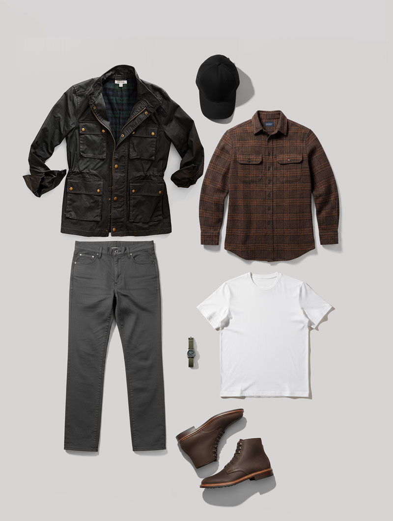 A flat lay display of a men’s smart casual outfit arranged on a light gray background. The items include a black utility jacket with gold snap buttons and a green plaid lining, a black baseball cap, a brown plaid flannel shirt, a plain white t-shirt, a pair of dark gray jeans, a silver wristwatch with a black face, and dark brown leather lace-up boots.