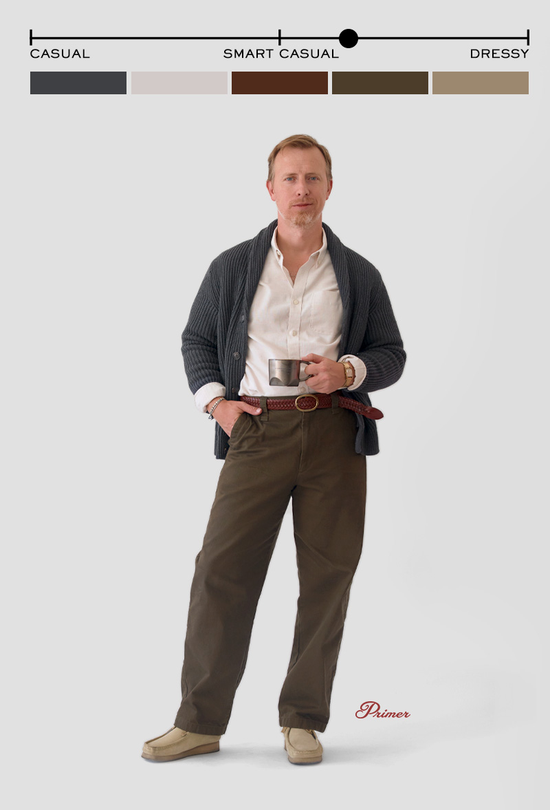 A man stands against a light grey background wearing a smart casual outfit. He has on a white button-down shirt partially tucked into olive green trousers with a braided brown leather belt. Over the shirt, he wears a chunky dark grey shawl-collar cardigan. His shoes are light beige suede moccasin-style loafers. He holds a metal cup in his right hand and wears a wristwatch and beaded bracelet on his left wrist. At the top of the image, a style scale ranging from “Casual” to “Dressy” marks the outfit as smart casual, accompanied by color swatches matching the clothing. The word “Primer” is written in red script near his feet.