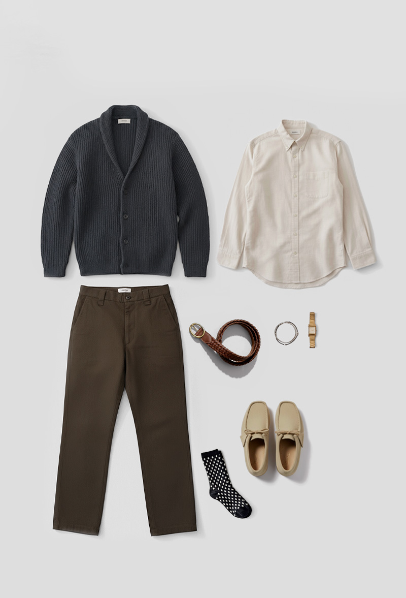 A flat lay of a smart casual men's outfit arranged on a light grey background. The ensemble includes a dark grey ribbed shawl-collar cardigan, an off-white button-down shirt, and a pair of olive green chinos. Accessories include a braided brown leather belt, a silver bracelet, a gold-toned wristwatch, and a pair of black socks with white polka dots. The shoes are light beige suede moccasin-style loafers.