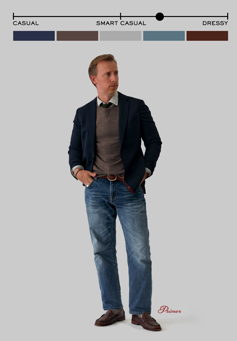 A man stands against a plain gray background wearing a smart casual outfit. He has on a navy blazer over a brown crew neck sweater and a light gray dress shirt with a green tie. He pairs this with medium-wash blue jeans, a red woven belt, and brown leather loafers. His hands are in his pockets, and he is looking slightly to the side. Above him is a style spectrum labeled “Casual” to “Dressy,” with a marker positioned under “Smart Casual.” Color swatches run beneath the spectrum, corresponding to outfit tones. The word “Primer” appears in red script near his feet.
