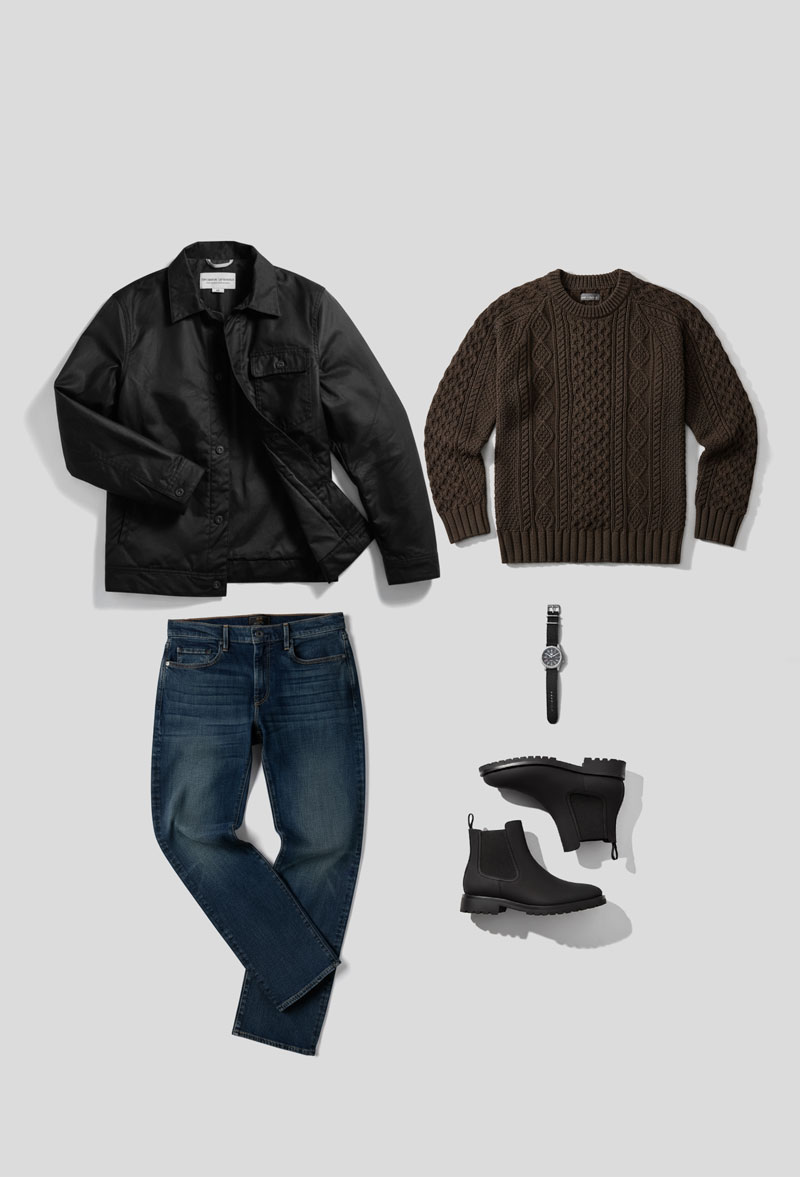 Flat lay of a smart casual men's outfit arranged on a light gray background. The ensemble includes a black waxed canvas button-up jacket from Thursday Boot Co with a collar in the top left, a brown cable-knit crew neck sweater in the top right, a pair of medium-wash blue jeans folded at the knees in the bottom left, a black leather-strapped wristwatch in the center, and a pair of black Chelsea boots with rugged soles positioned in the bottom right.
