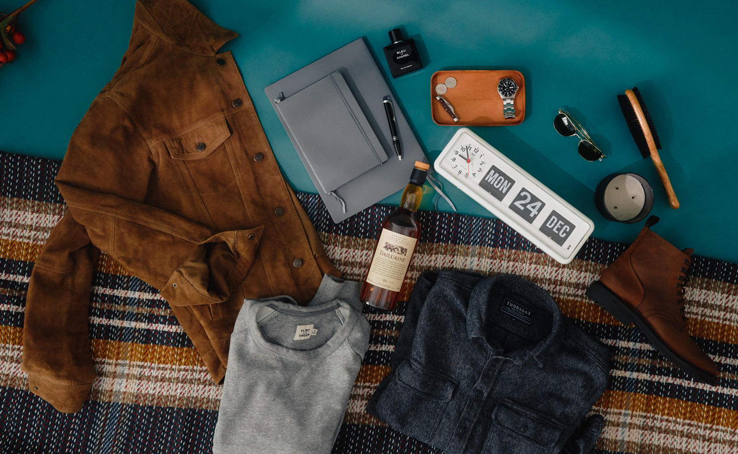 Flat lay image showcasing a men’s holiday gift guide with curated gift ideas arranged on a blue surface and plaid blanket. Items include a brown suede jacket, folded grey sweatshirt, folded dark grey button-up shirt, brown leather boot, bottle of Dailuaine 16-year-old Scotch whisky, two grey notebooks with a black pen, black bottle of Bleu de Chanel cologne, tan leather valet tray holding a wristwatch, coins, and a key, aviator sunglasses, wooden hairbrush, round candle, and a retro-style flip clock