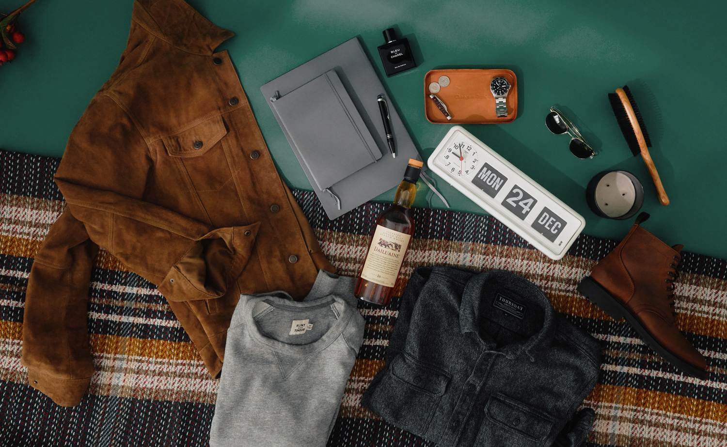 Flat lay image showcasing a men’s holiday gift guide with curated gift ideas arranged on a blue surface and plaid blanket. Items include a brown suede jacket, folded grey sweatshirt, folded dark grey button-up shirt, brown leather boot, bottle of Dailuaine 16-year-old Scotch whisky, two grey notebooks with a black pen, black bottle of Bleu de Chanel cologne, tan leather valet tray holding a wristwatch, coins, and a key, aviator sunglasses, wooden hairbrush, round candle, and a retro-style flip clock