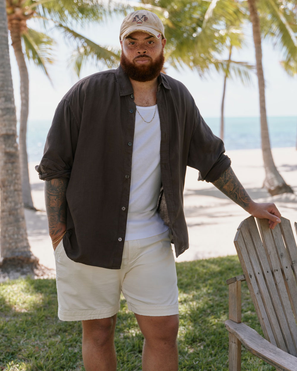 Man at the beach wearing a white ribbed tank top under an open dark brown linen shirt, with off-white shorts and a tan baseball cap