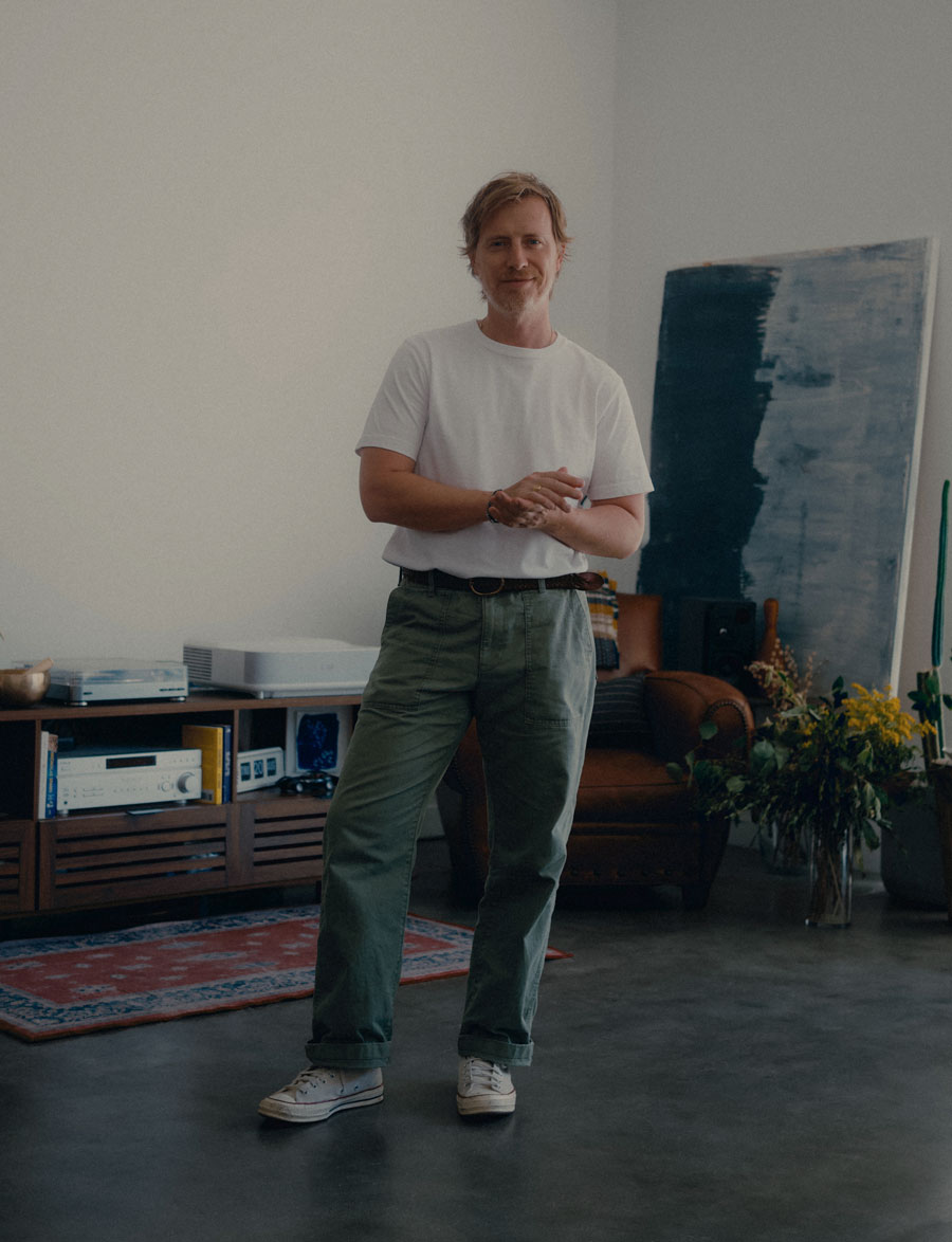 Andrew wearing a men's outfit featuring free assembly utility pants that are an homage to OG 107 Fatigue pants. he wears them with a tucked in white tshirt and beige tan parchment color converse high tops