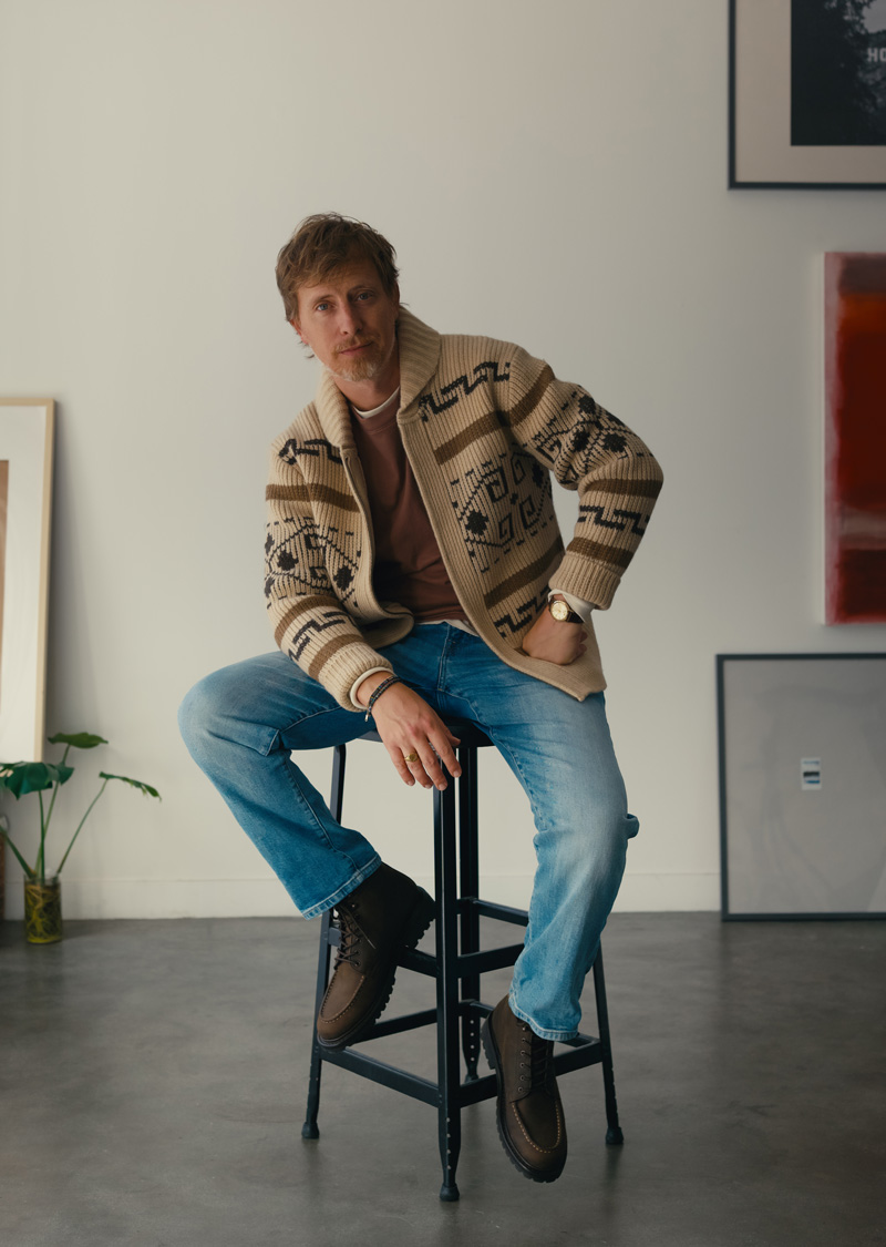Man seated on a stool in a tan Pendleton Westerley cardigan, faded red tee, light wash jeans and brown moc toe boots